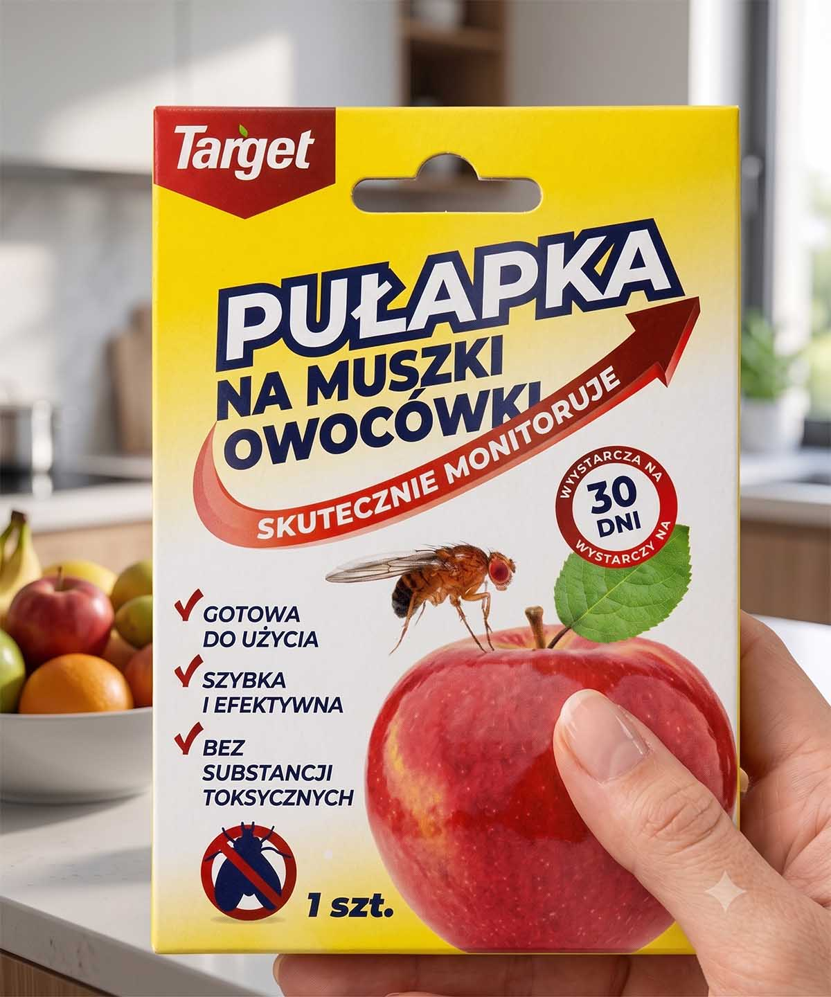 Kobieca dłoń trzymająca oryginalne, żółto-białe opakowanie blister pułapki na muszki owocówki Target z grafiką czerwonego jabłka.